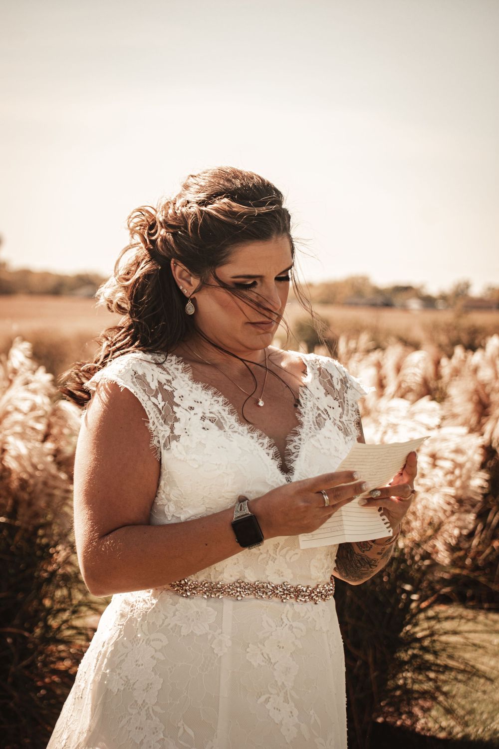 A bride in a lace white dress reads from a letter in a golden sunlit field with autumn pampas grass.