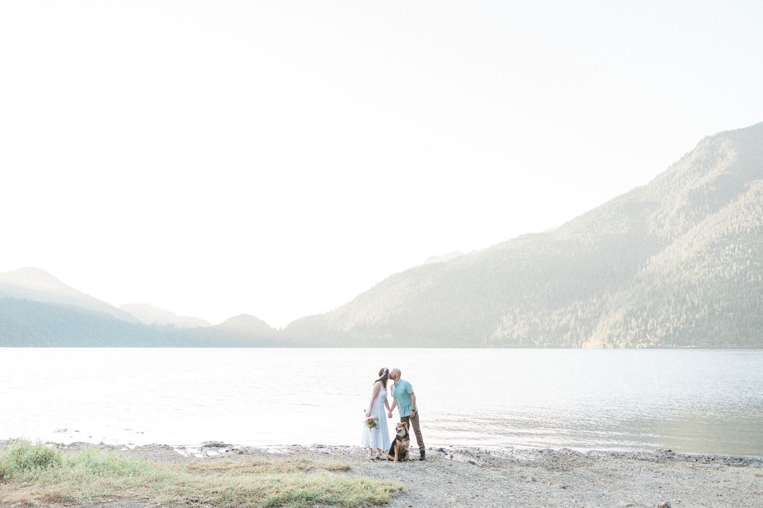 Two people stand together on a rocky beach with mountains and water in the background during golden hour at sunrise.