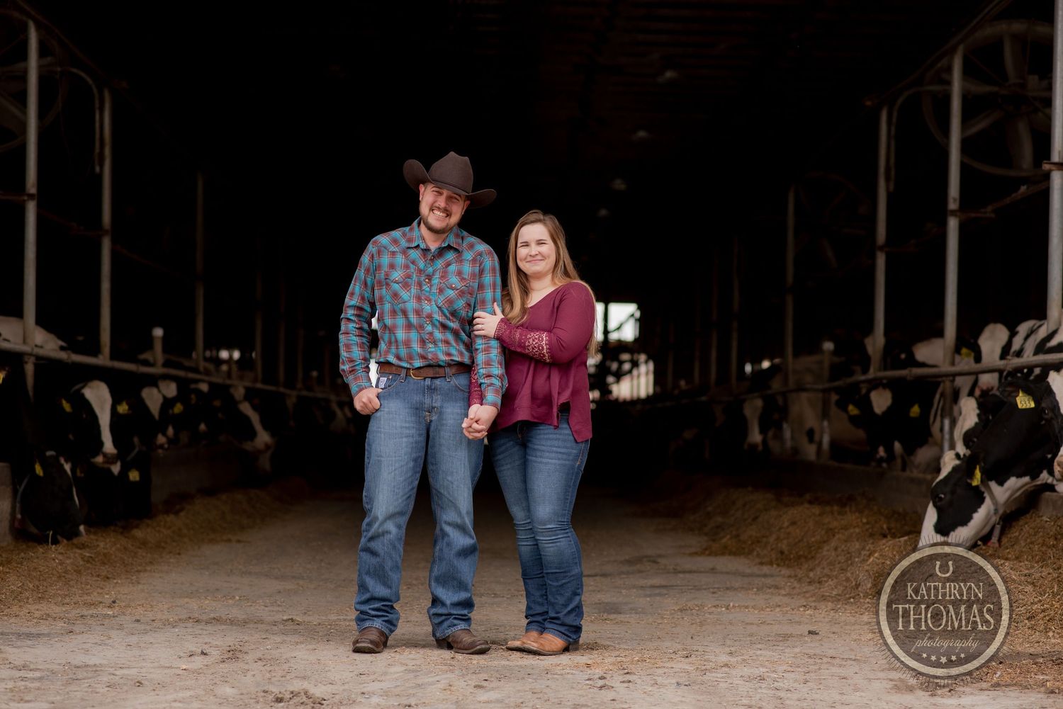 Couple posing together in western attire inside a dairy barn with cattle visible.
