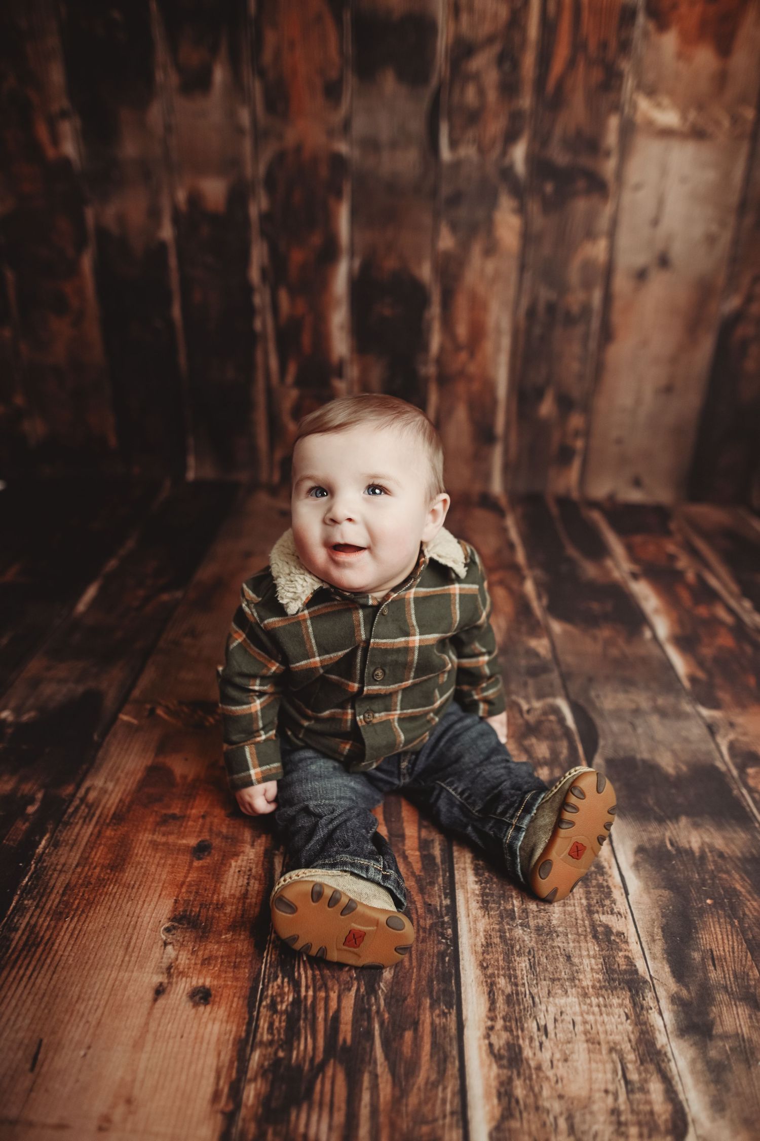 Baby in plaid shirt sitting on rustic wooden floor with vintage backdrop.