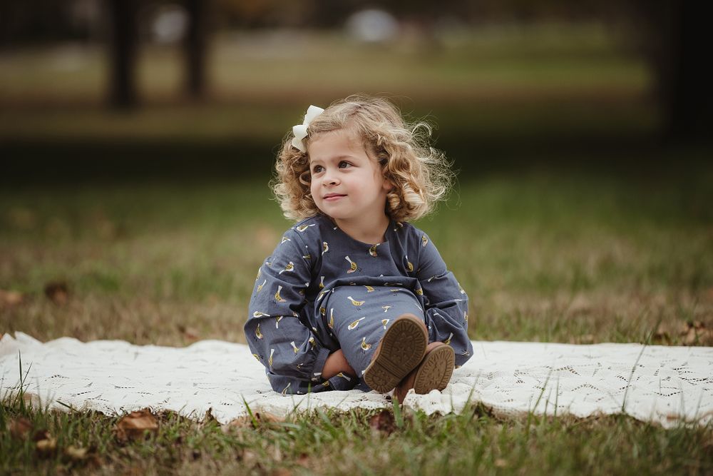 A toddler in a blue dress sits on a white blanket during an outdoor autumn photoshoot.