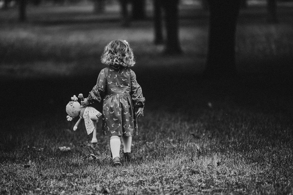 A black and white photo of a small child walking through a park holding a stuffed toy.