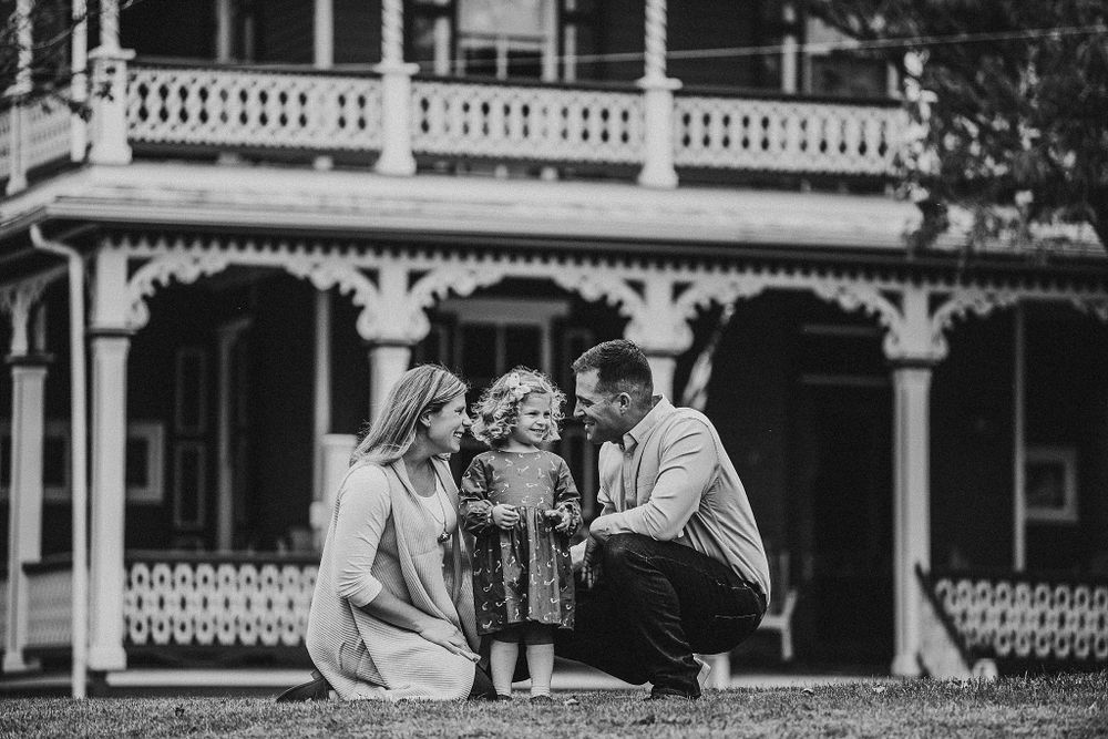 Black and white portrait of family sharing intimate moment in front of Victorian mansion.