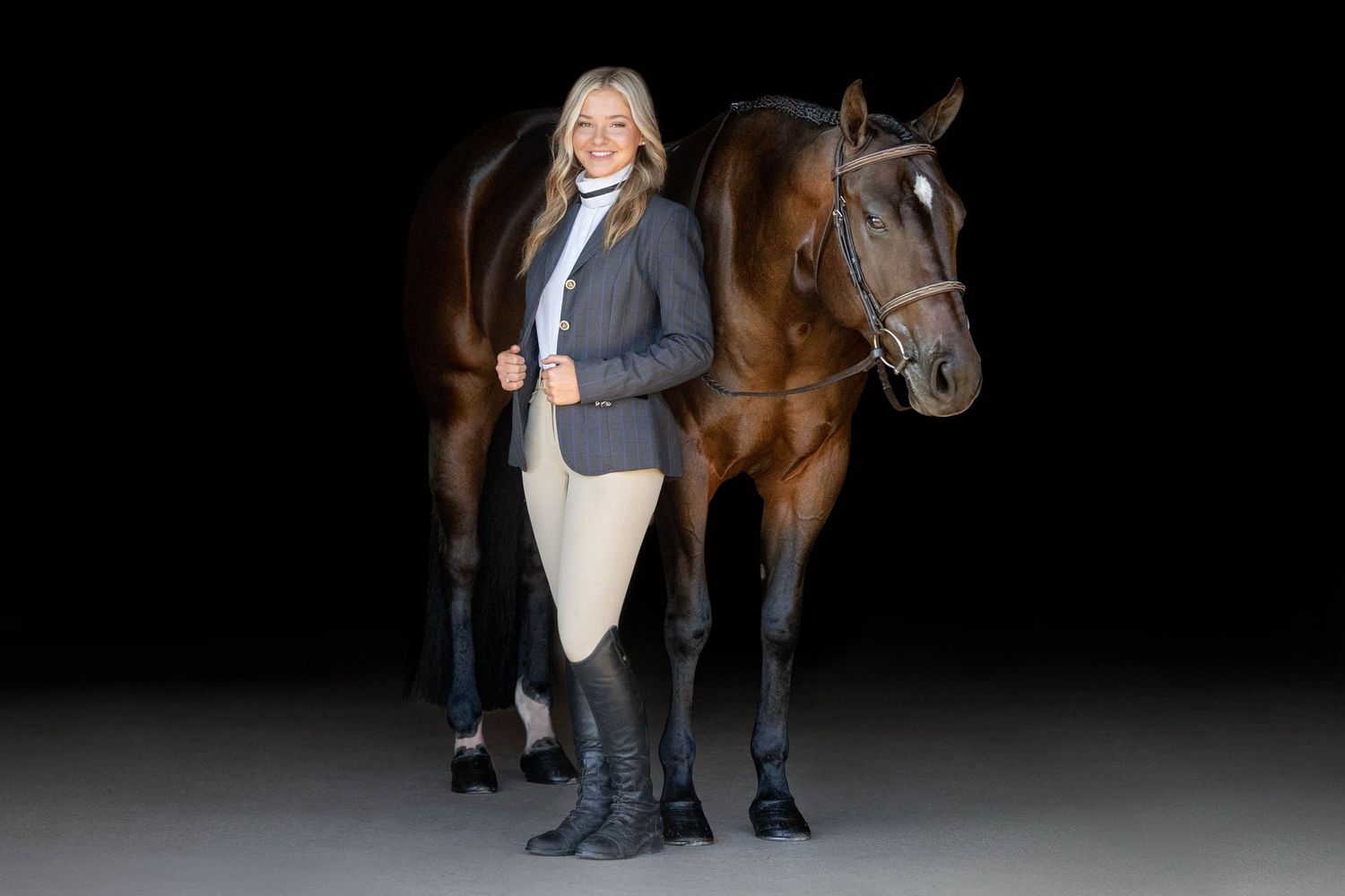 Black background image of a girl in huntseat coat and breeches standing next to bay mare in bridle
