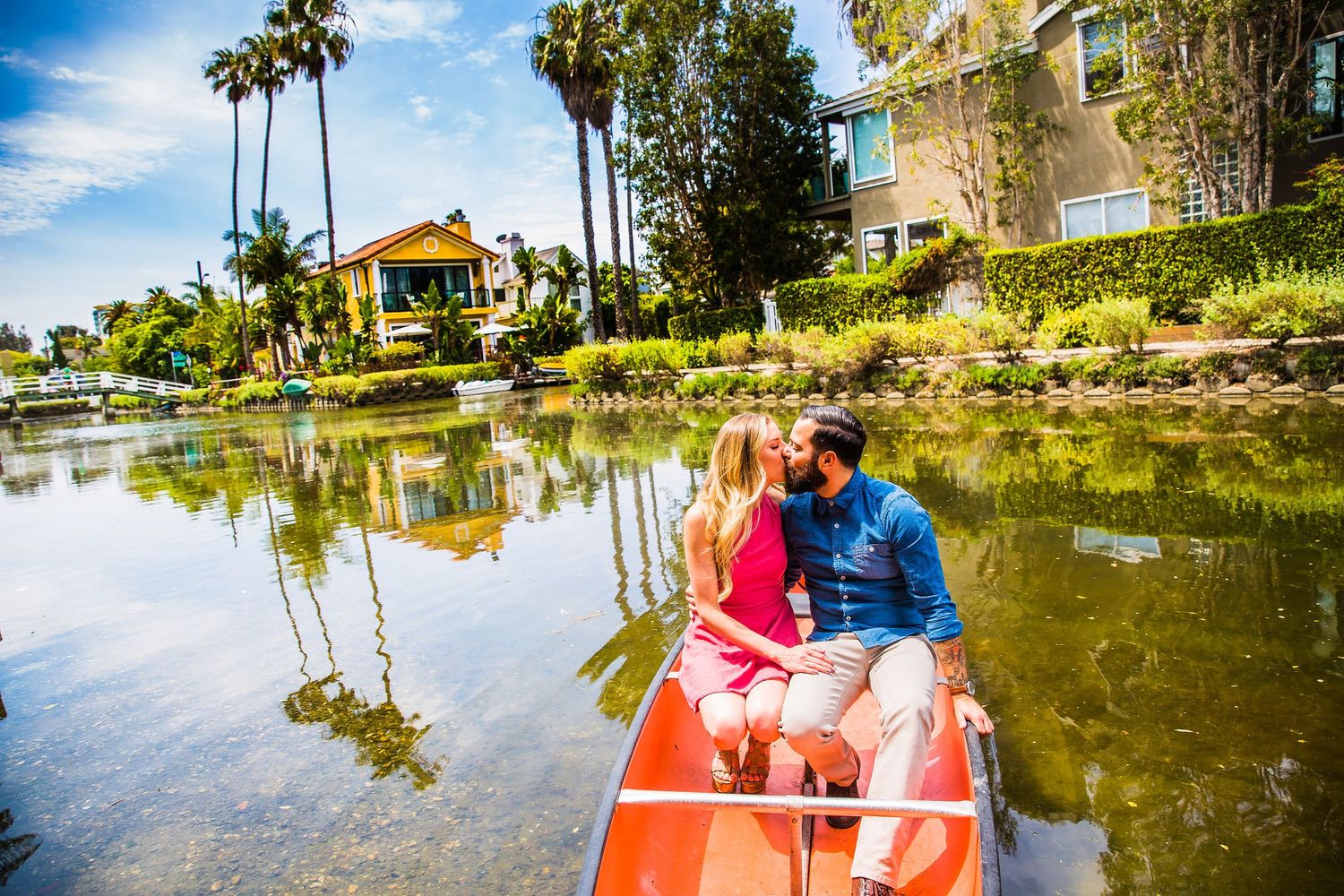 Couple sharing romantic moment in red canoe on lake with palm trees and luxury homes in background.