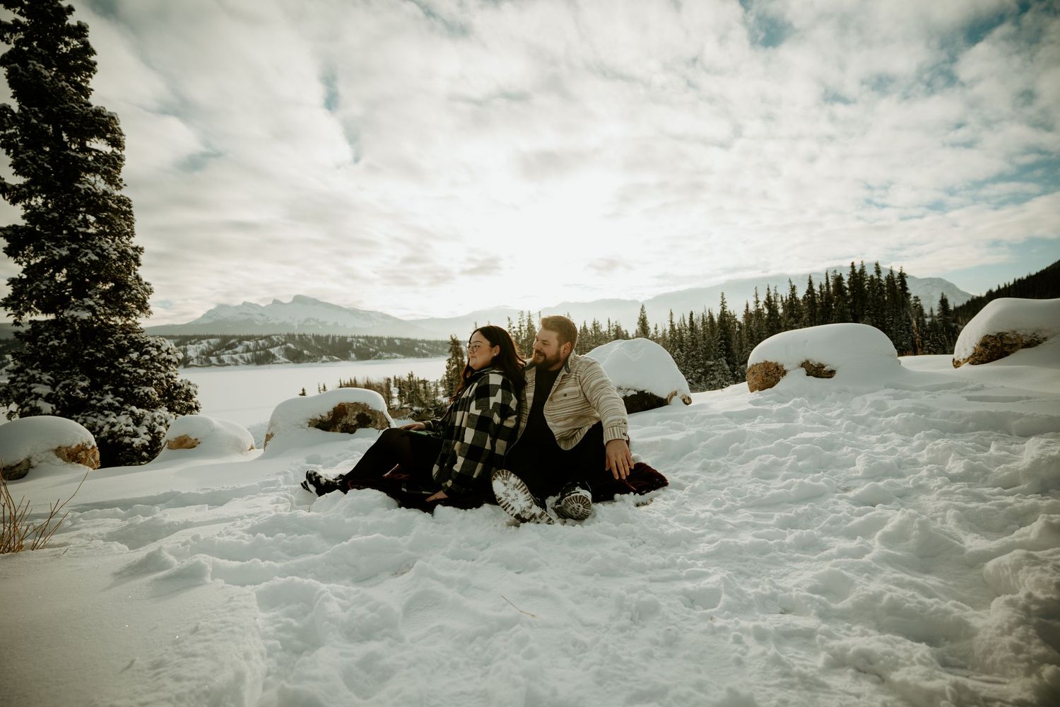 Couple sitting together in deep snow with mountains and evergreen forest in the background.