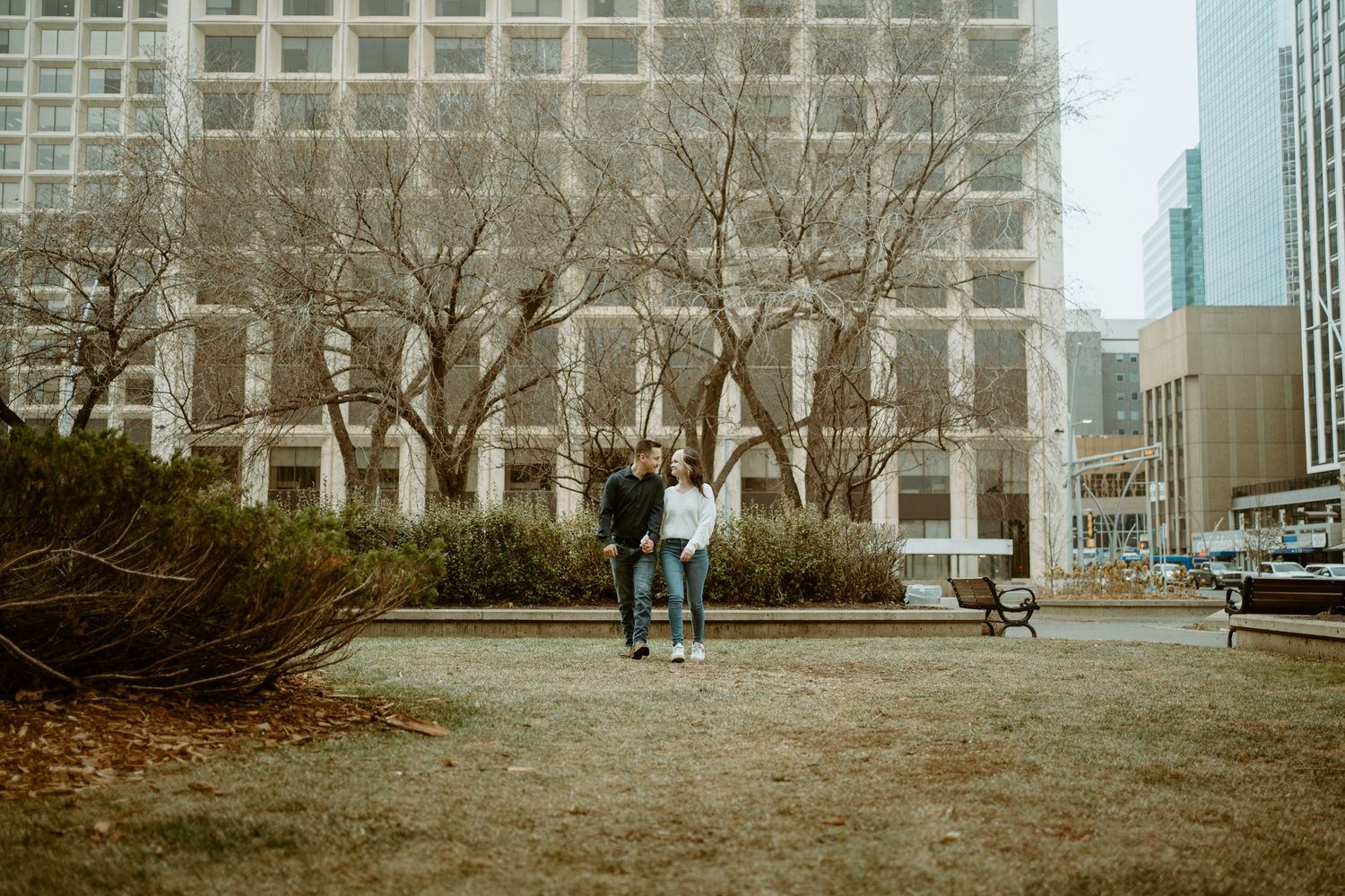 Distant shot of couple embracing in urban park with modernist building in background.