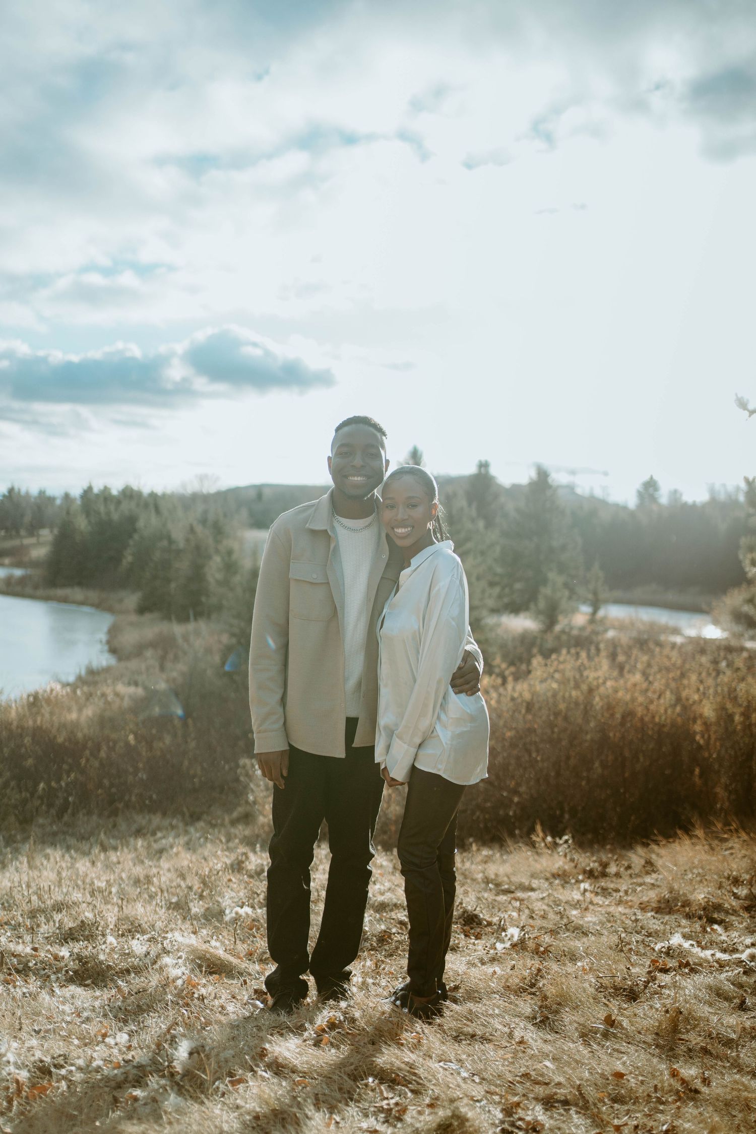 Couple standing close together on a hilltop overlooking a serene lake and forest landscape.