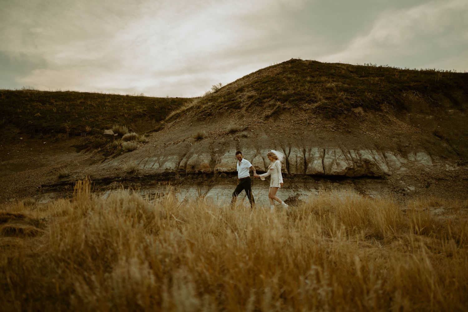 Elopement photos of a couple running through tall grass against a dramatic mountainous backdrop in sepia tones in Drumheller, Alberta.