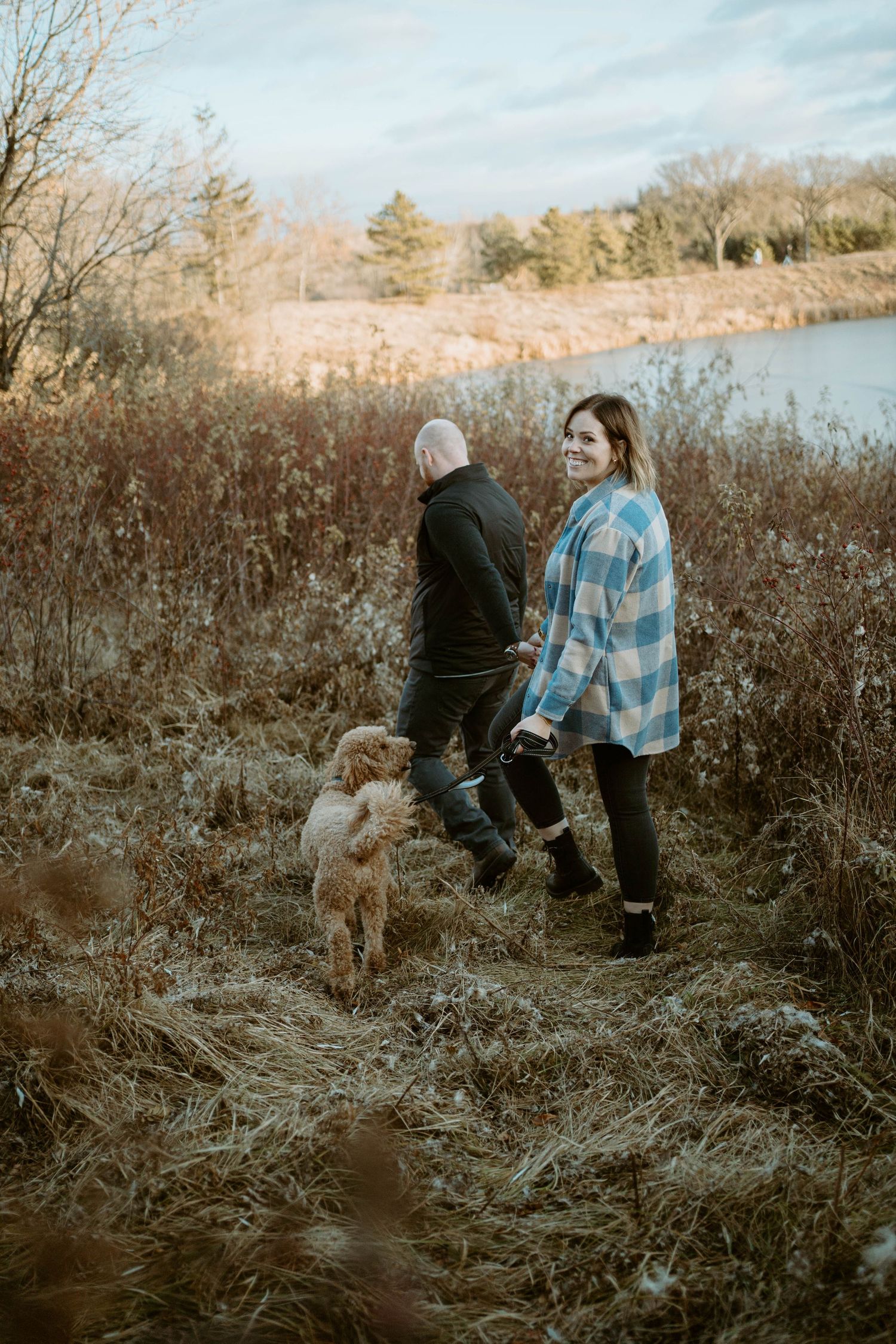 Two people walking with a dog along a lakeside trail during golden hour in autumn.