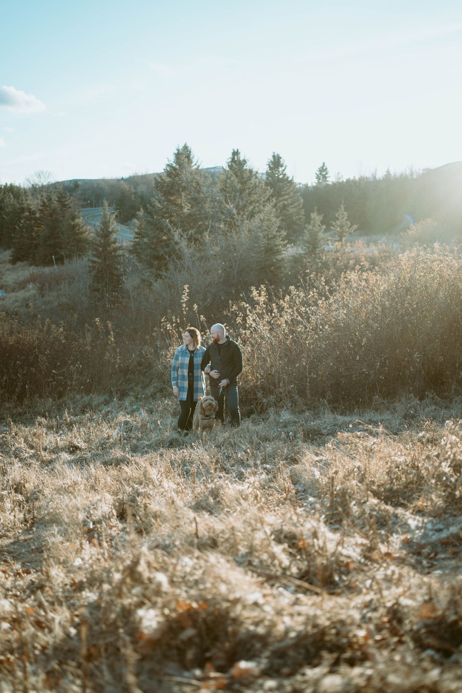 A couple stands together on a hillside with evergreen trees and a lake in the distance.
