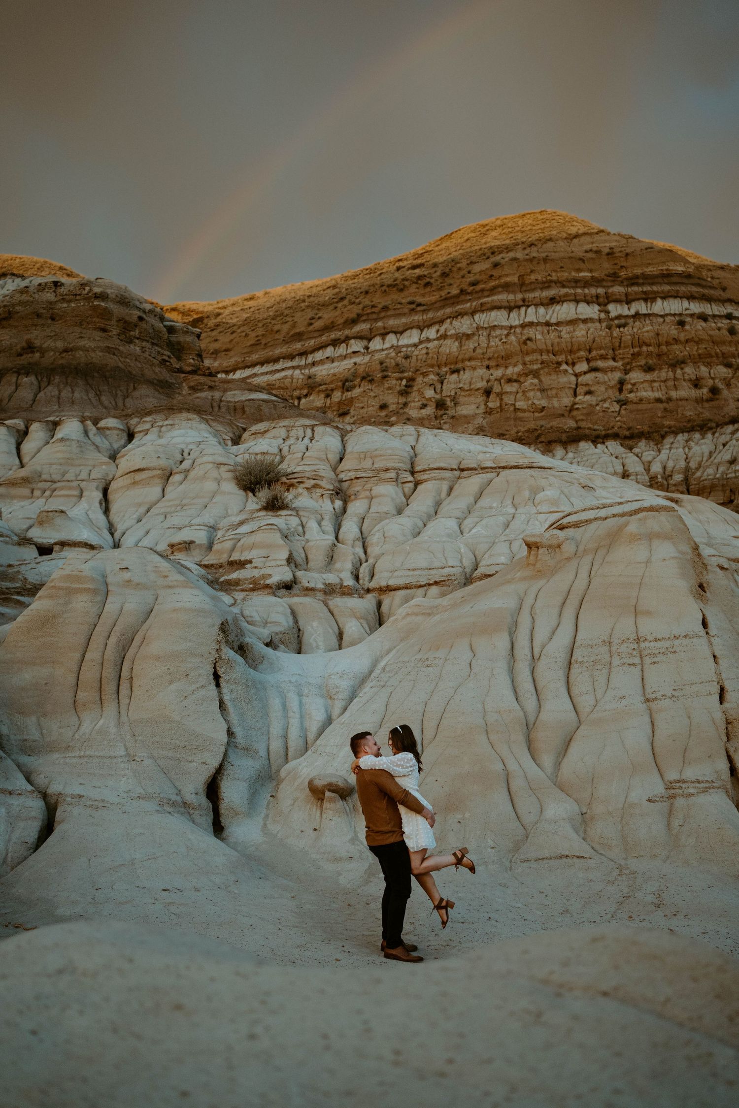 Romantic moment between couple against dramatic white rock formations with rainbow in background.