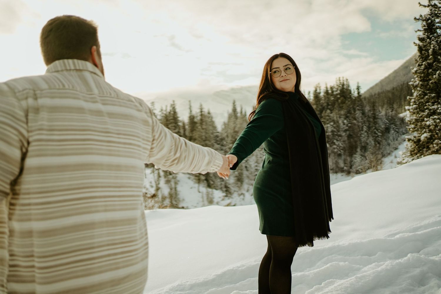 A couple enjoys a playful moment in a snowy winter landscape surrounded by evergreen trees and mountains.