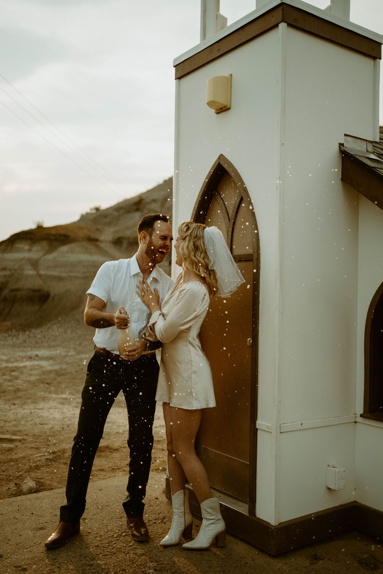 Couple shares intimate moment outside small white chapel door with champagne falling in Drumheller, Alberta.