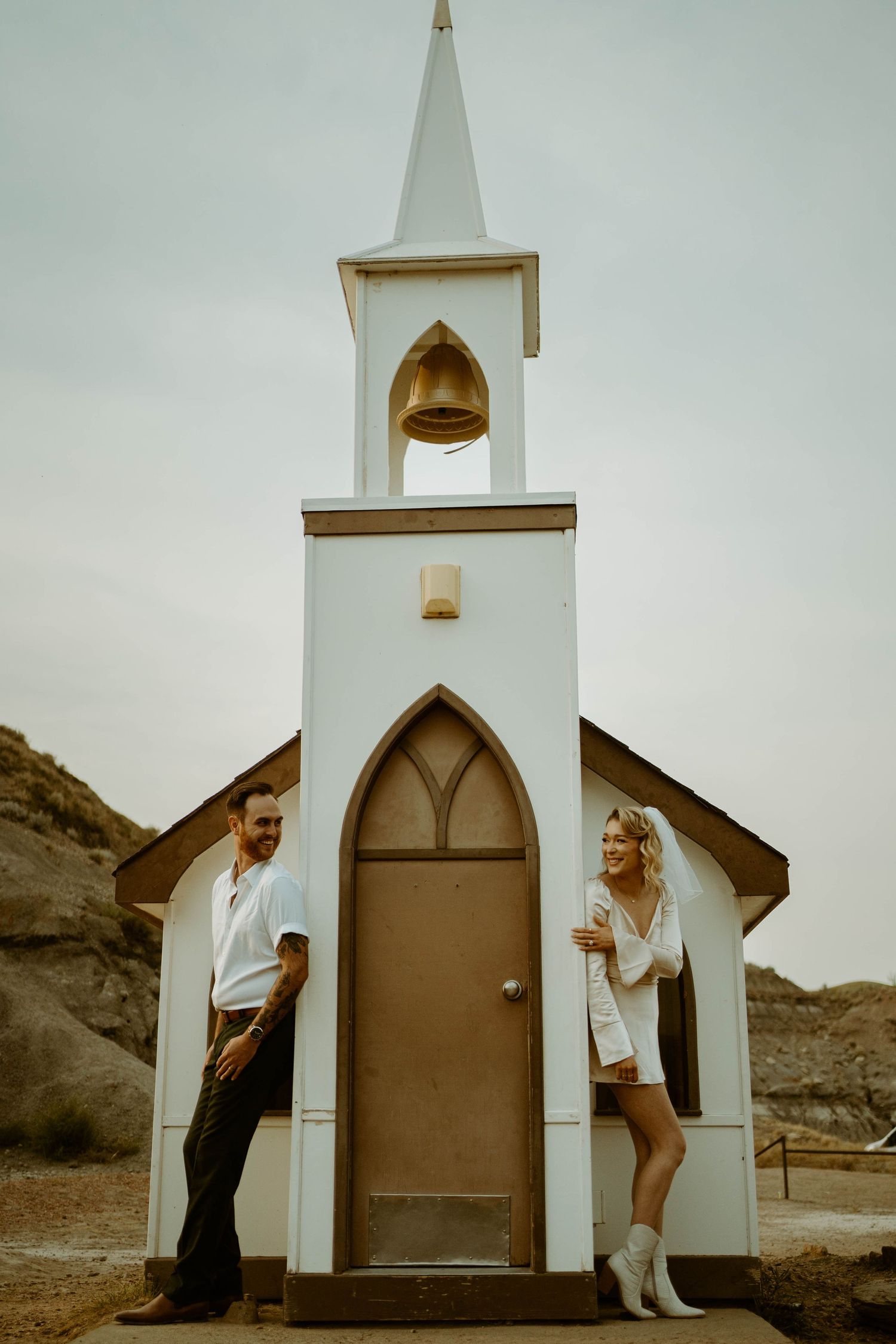 Couple poses playfully on either side of small white country church entrance in Drumheller, Alberta.