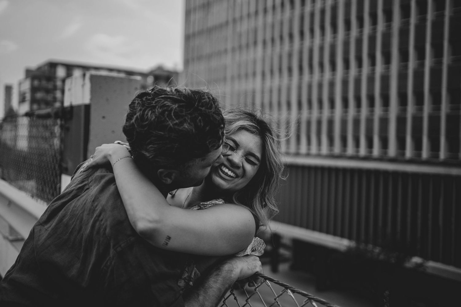 A romantic black and white photograph of a couple embracing and laughing on an urban rooftop.