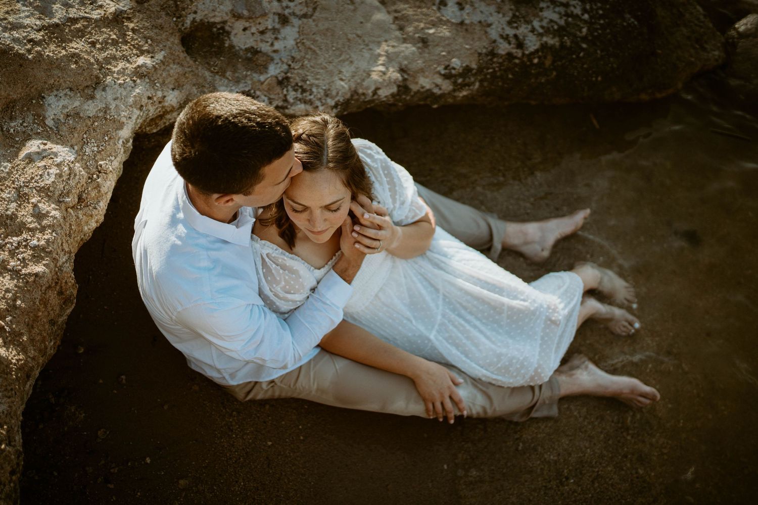 Intimate couple portrait on rocky beach in white attire during sunset.