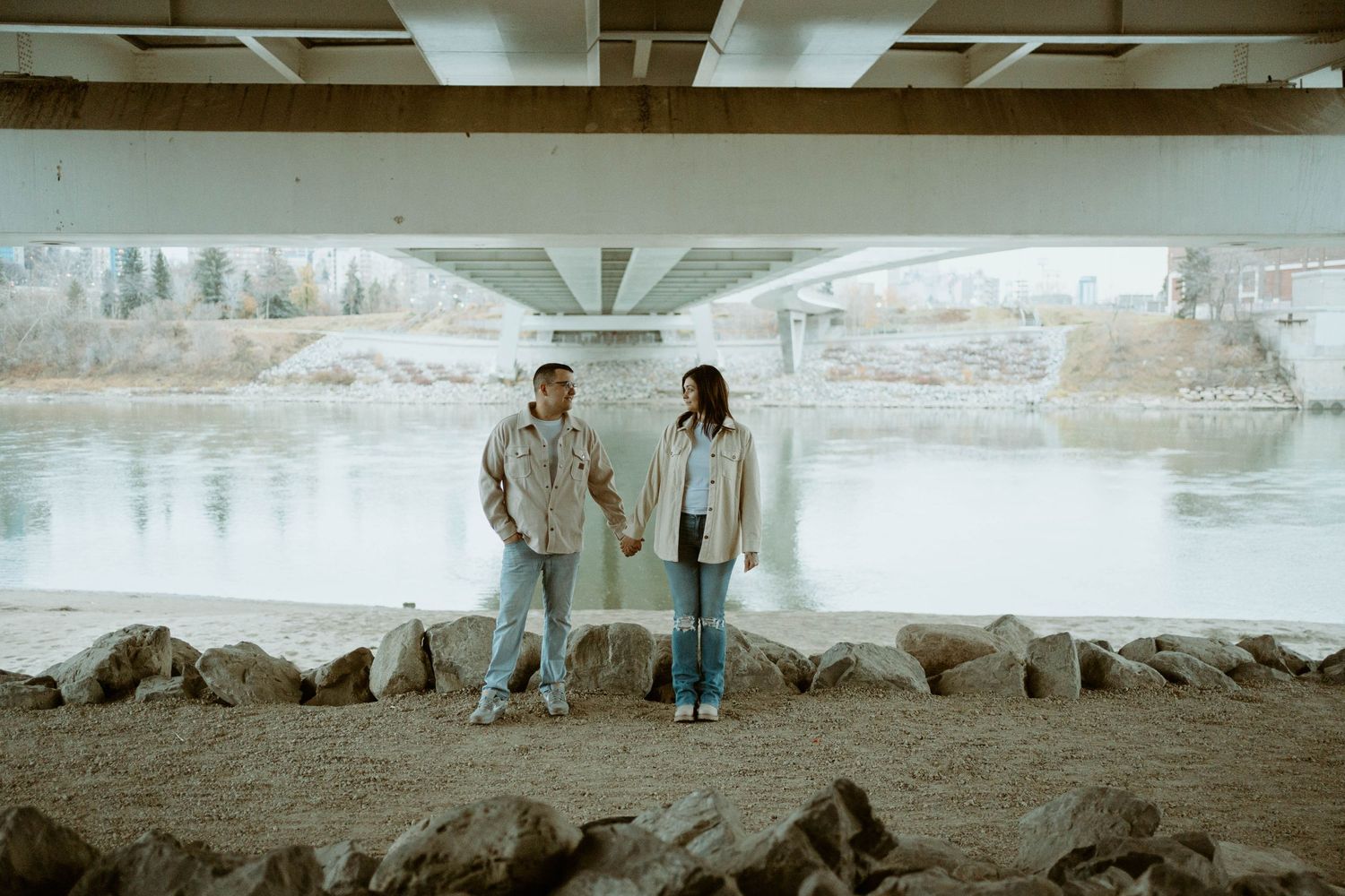 Couple holding hands while standing under a concrete bridge near rocky riverbank at sunset in Edmonton, Alberta.