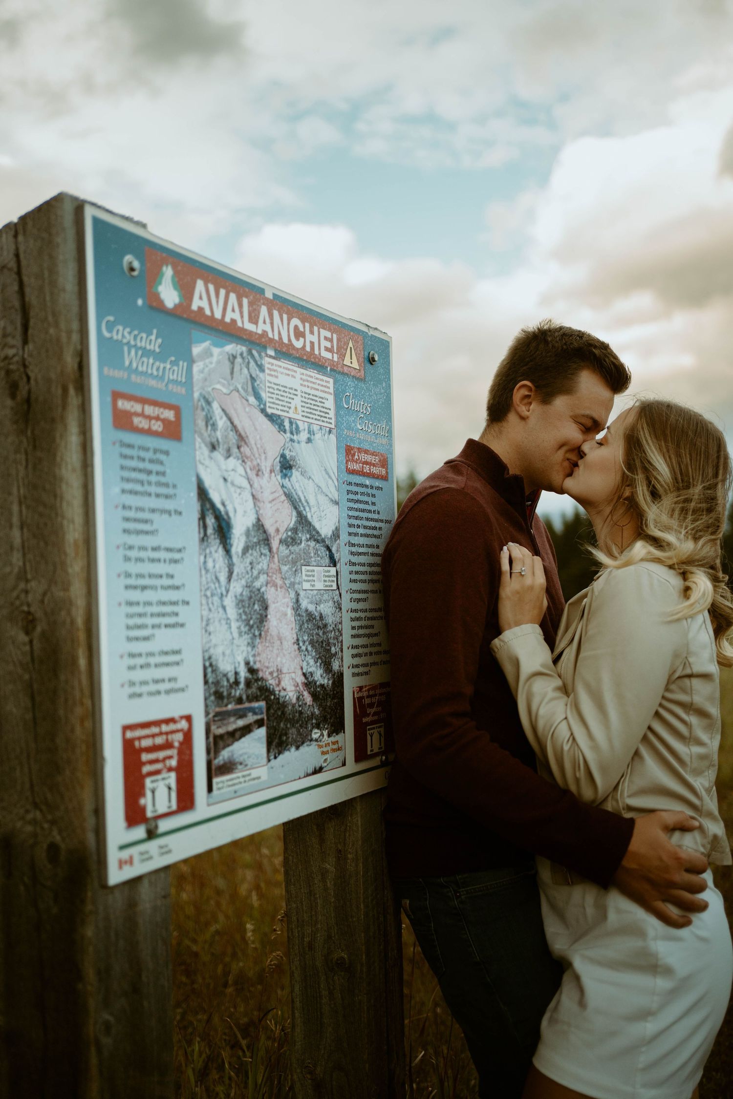 Couple embraces next to Avalanche Area warning sign in mountain setting in Banff, Alberta for an engagement session.