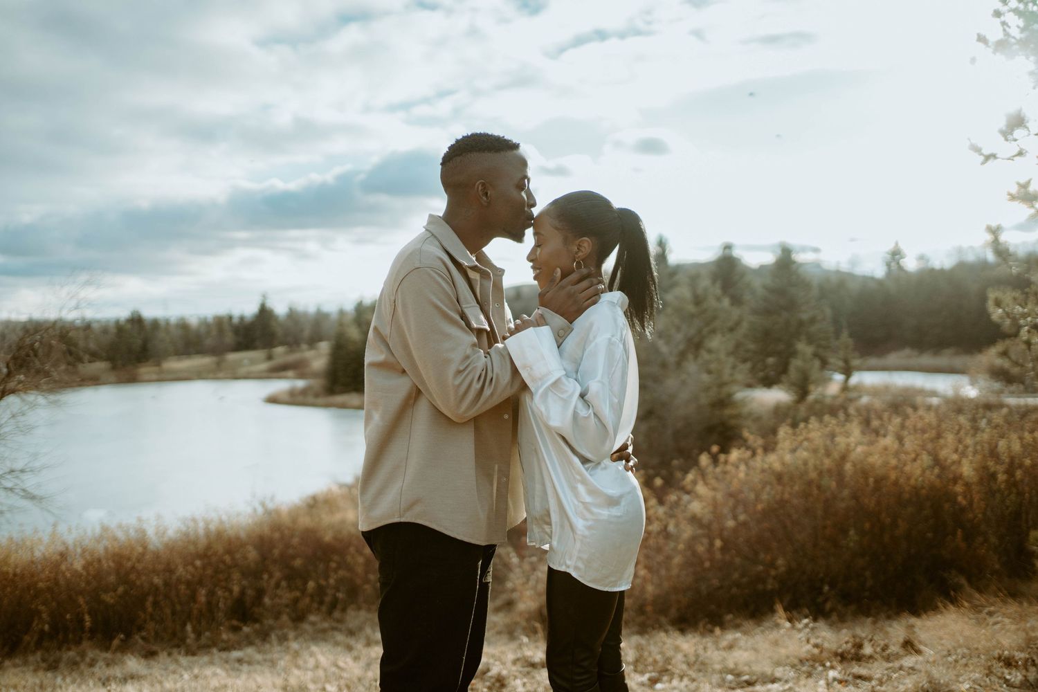 Intimate outdoor portrait of a couple embracing by a lake with autumn foliage in the background.