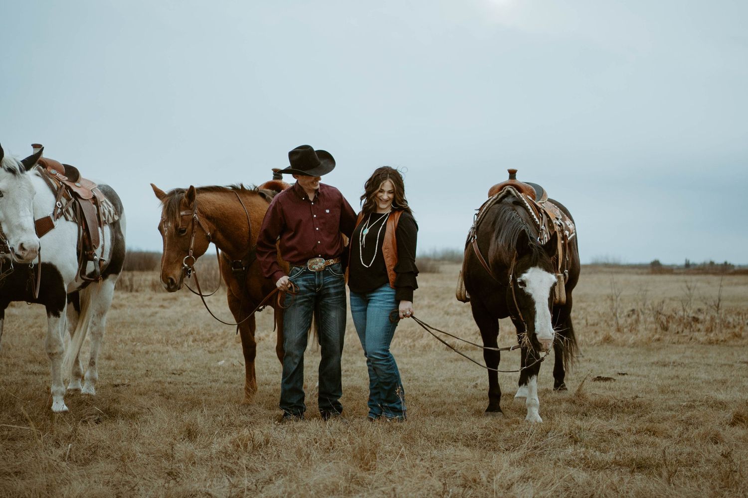 A couple stands between horses on a rural plain during an overcast day.