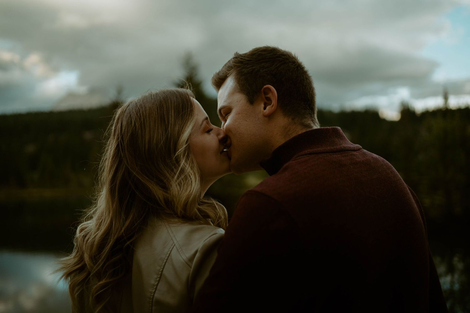 Romantic couple shares intimate kiss against cloudy mountain landscape in Banff, Alberta for an engagement session.