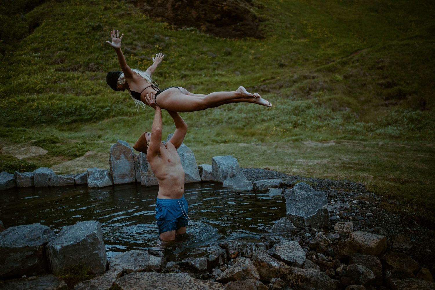 A dramatic lift pose performed in a natural hot spring surrounded by rocks in Iceland.