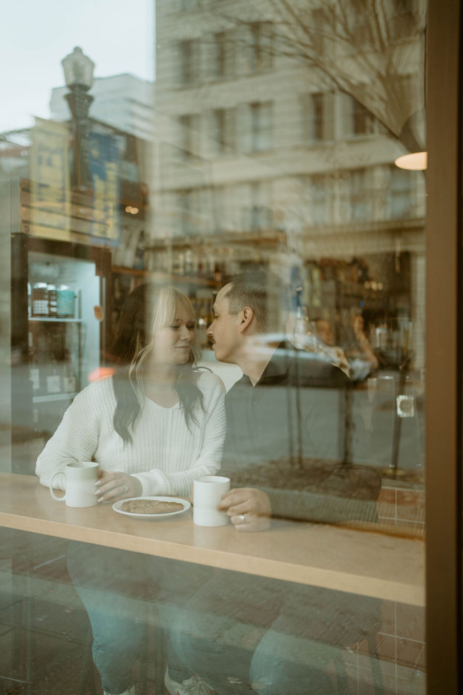 Couple enjoying coffee date by window in urban cafe with city reflection.
