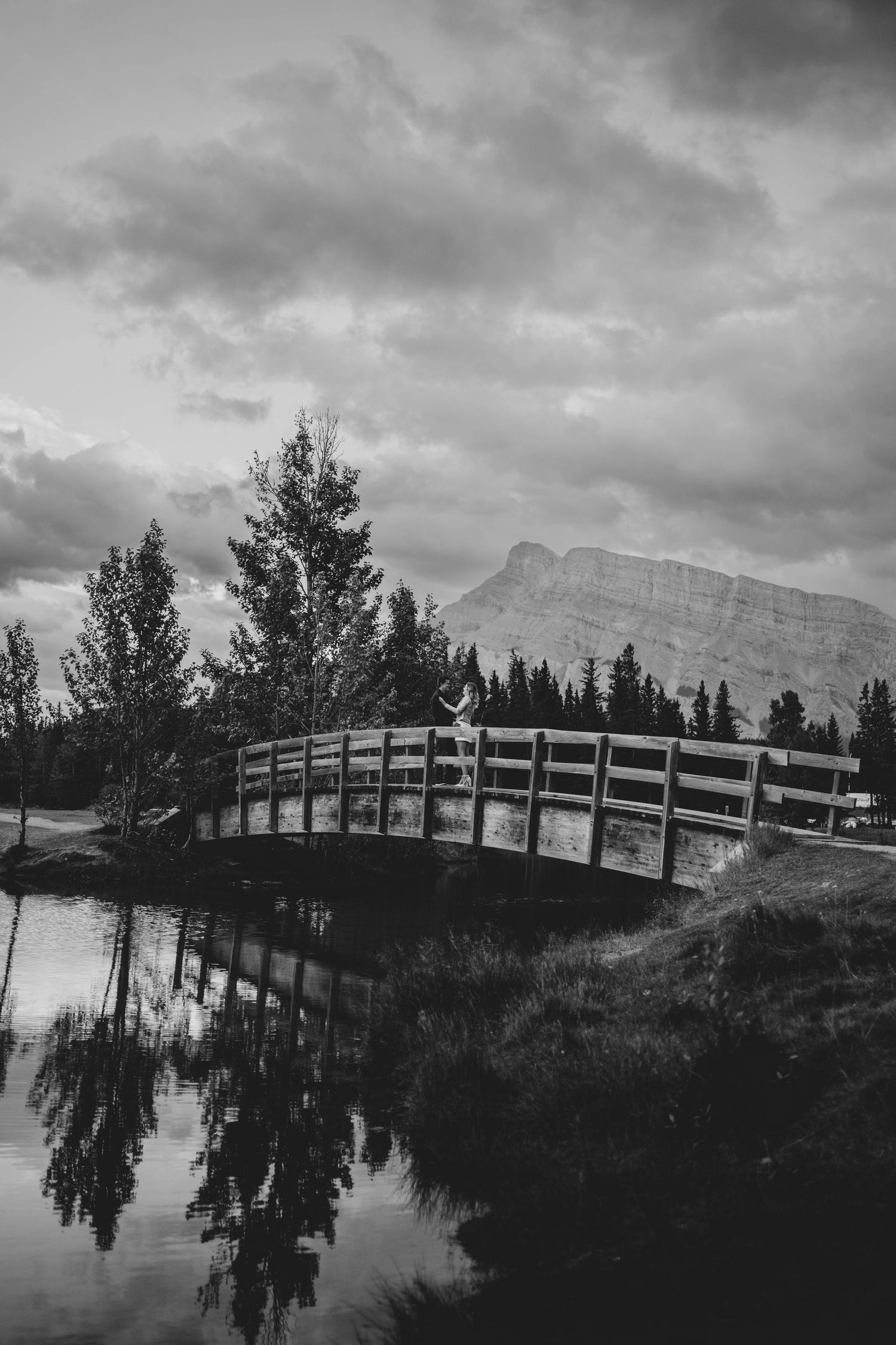 Black and white landscape with wooden bridge and couple over calm water reflecting mountain and cloudy sky in Banff, Alberta.