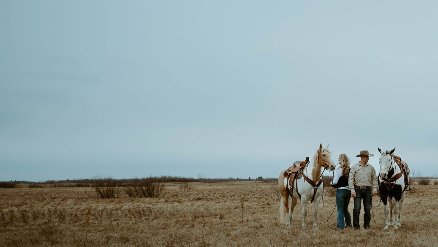 People walk with horses across an open field against a moody sky.