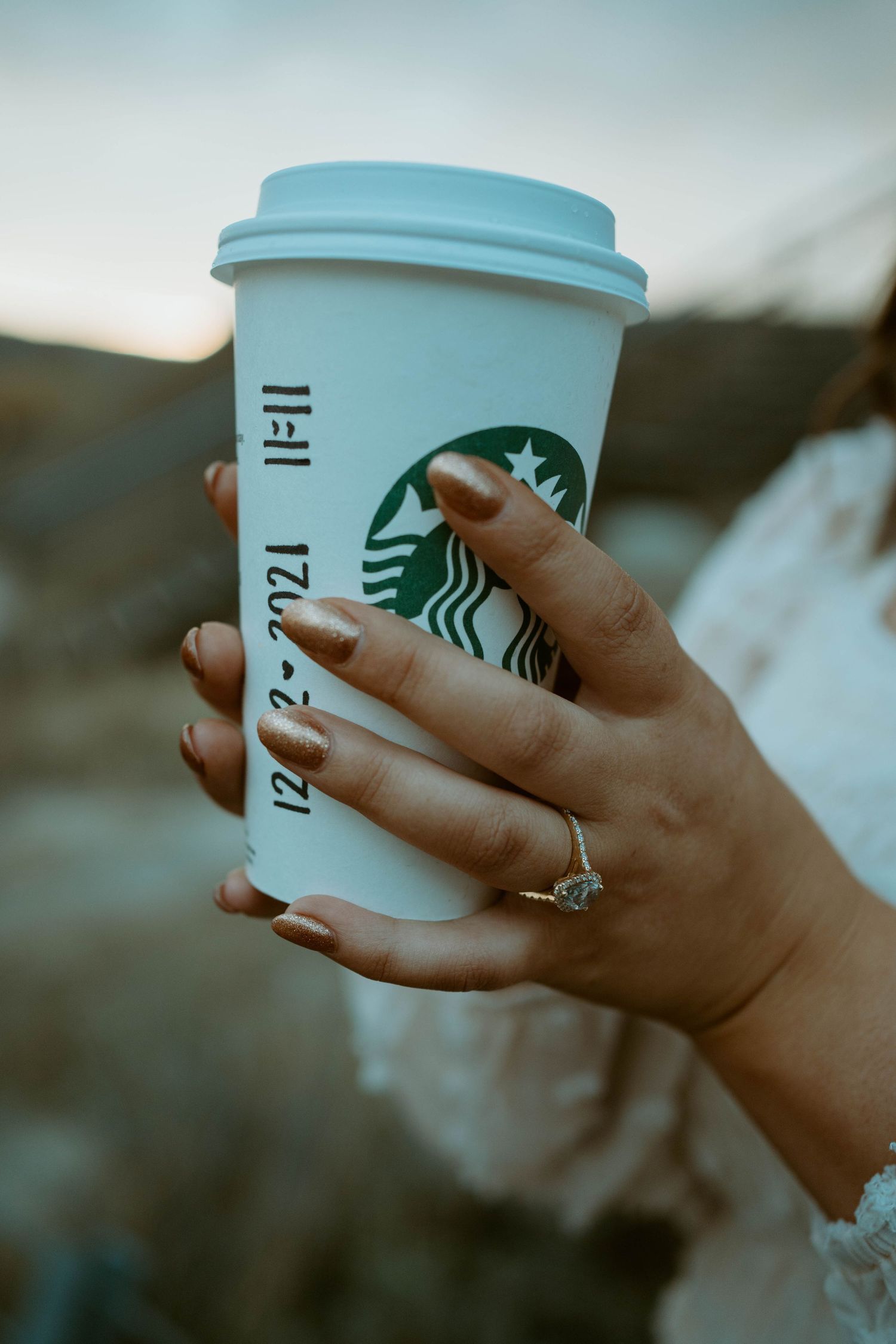 Hand with engagement ring holds a Starbucks coffee cup against sunset backdrop.