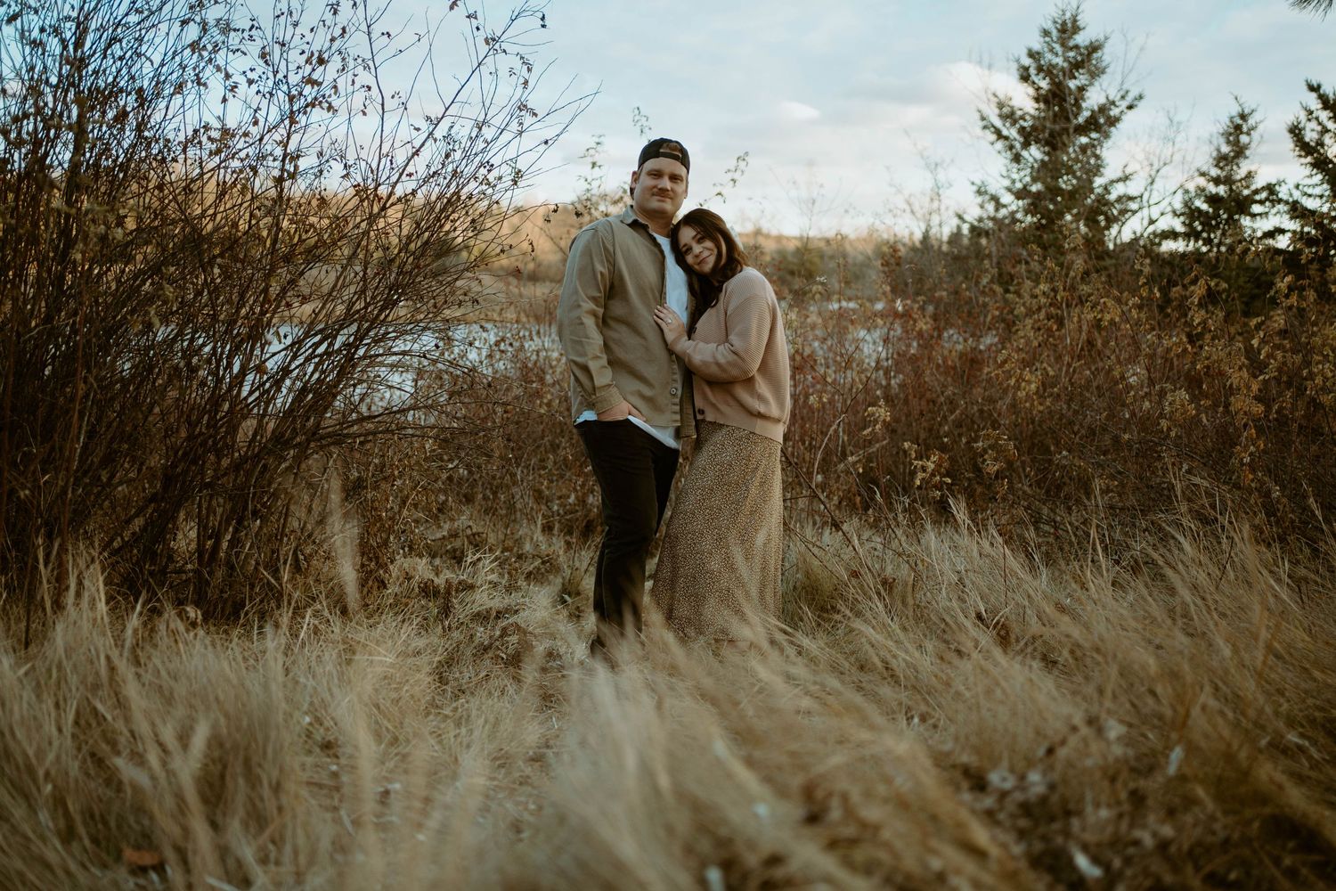 A couple stands together in tall grass with a lake view in the background.