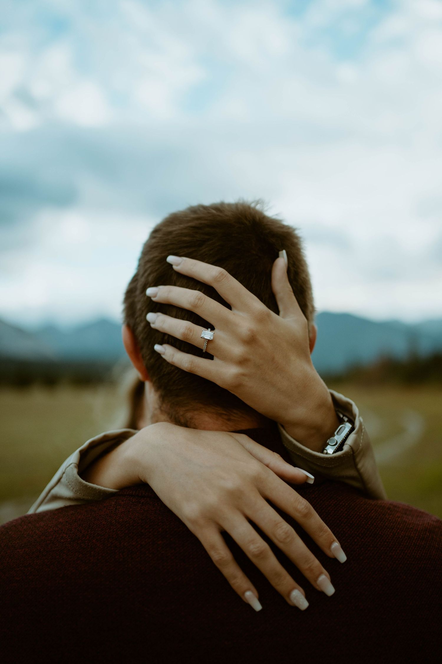 Hands wearing engagement ring embrace from behind against mountain backdrop in Banff, Alberta for an engagement session.