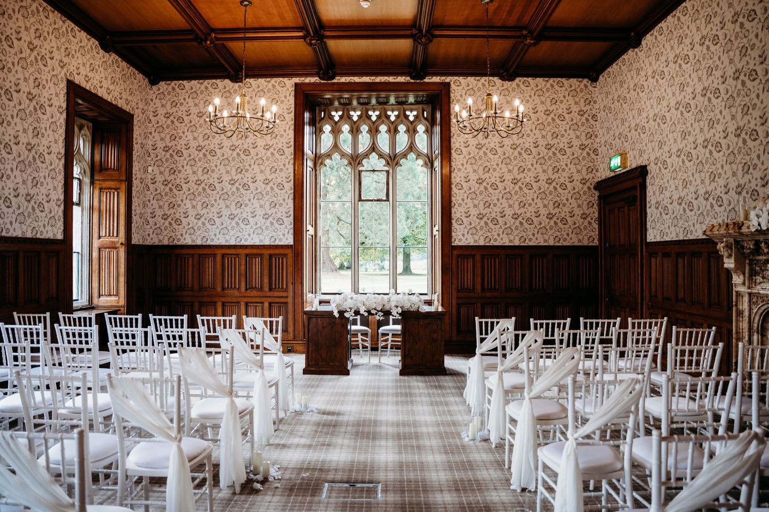 Traditional ceremony room with wooden paneling and white chairs facing gothic window.