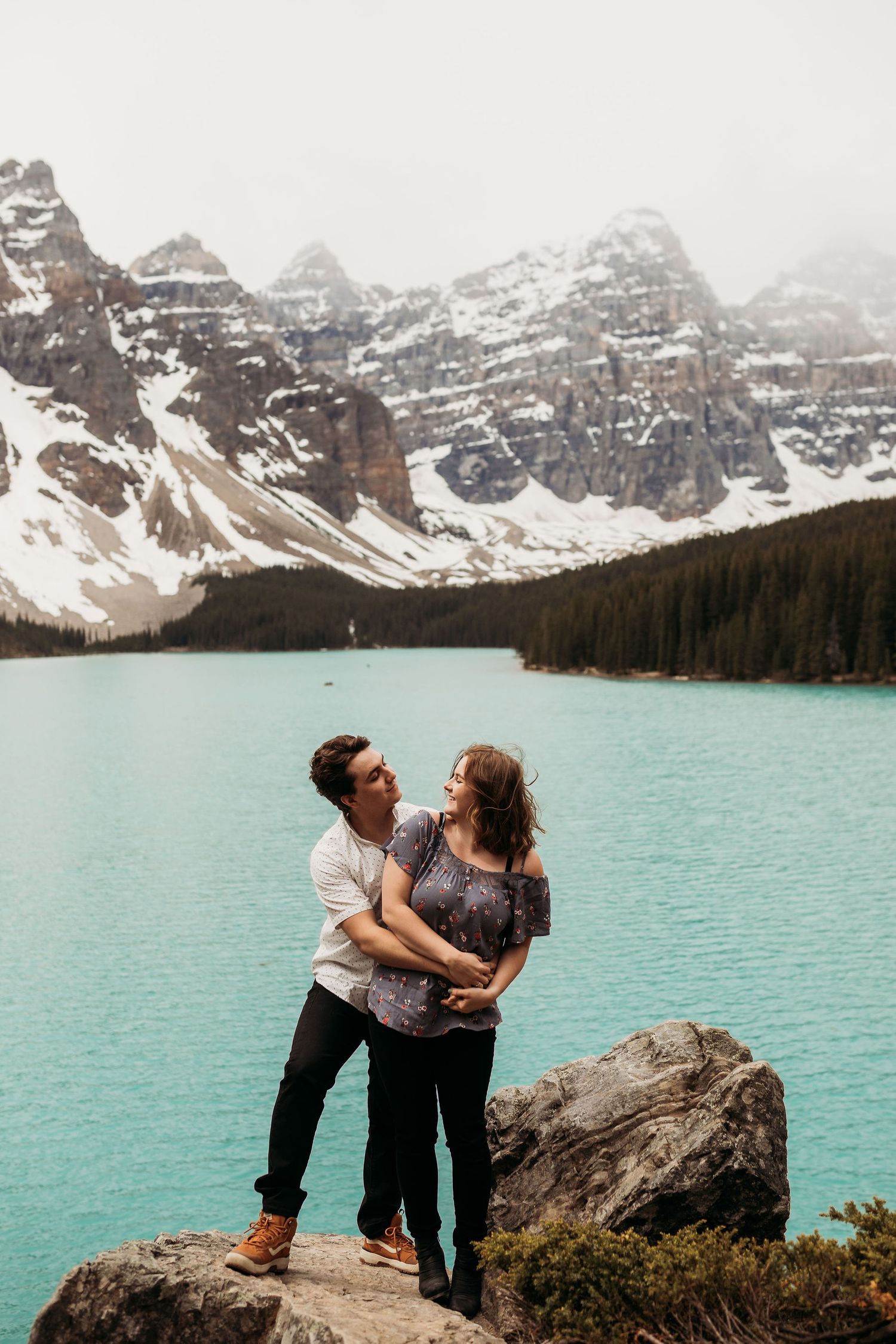 Romantic couple standing on rock formation at Moraine Lake with dramatic mountain backdrop.