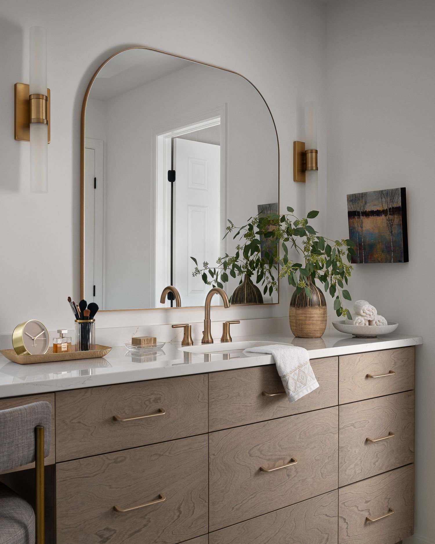 Modern bathroom vanity with arched mirror, brass fixtures, and wooden drawers featuring natural decor elements.