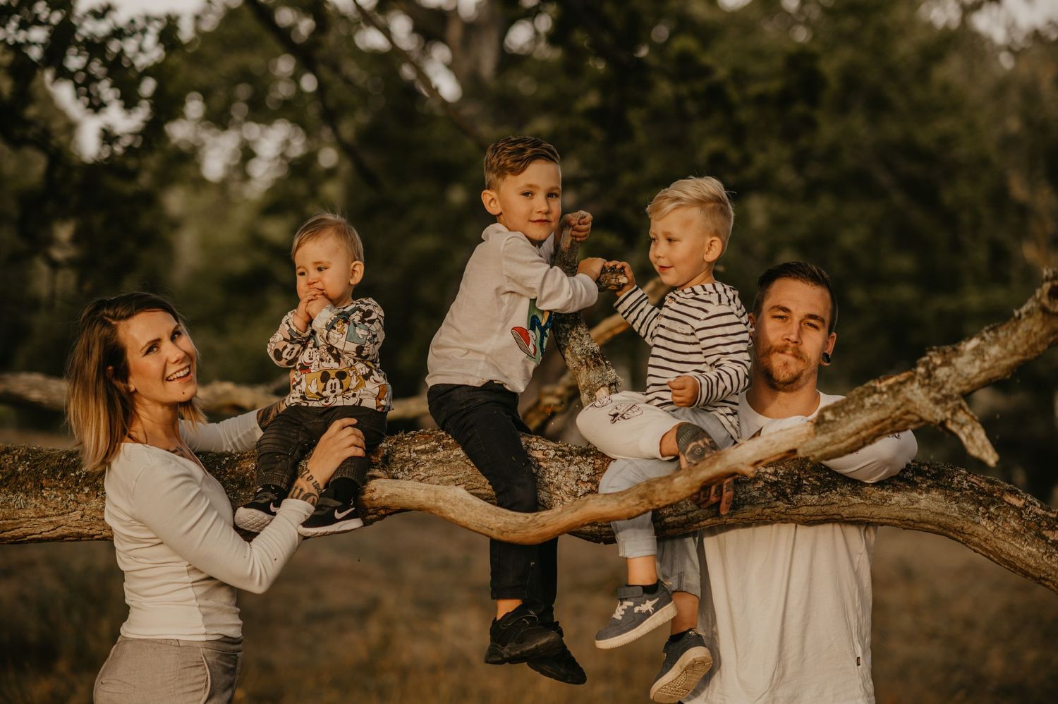 Happy family with three young children climbing a large tree branch outdoors in a wooded setting.