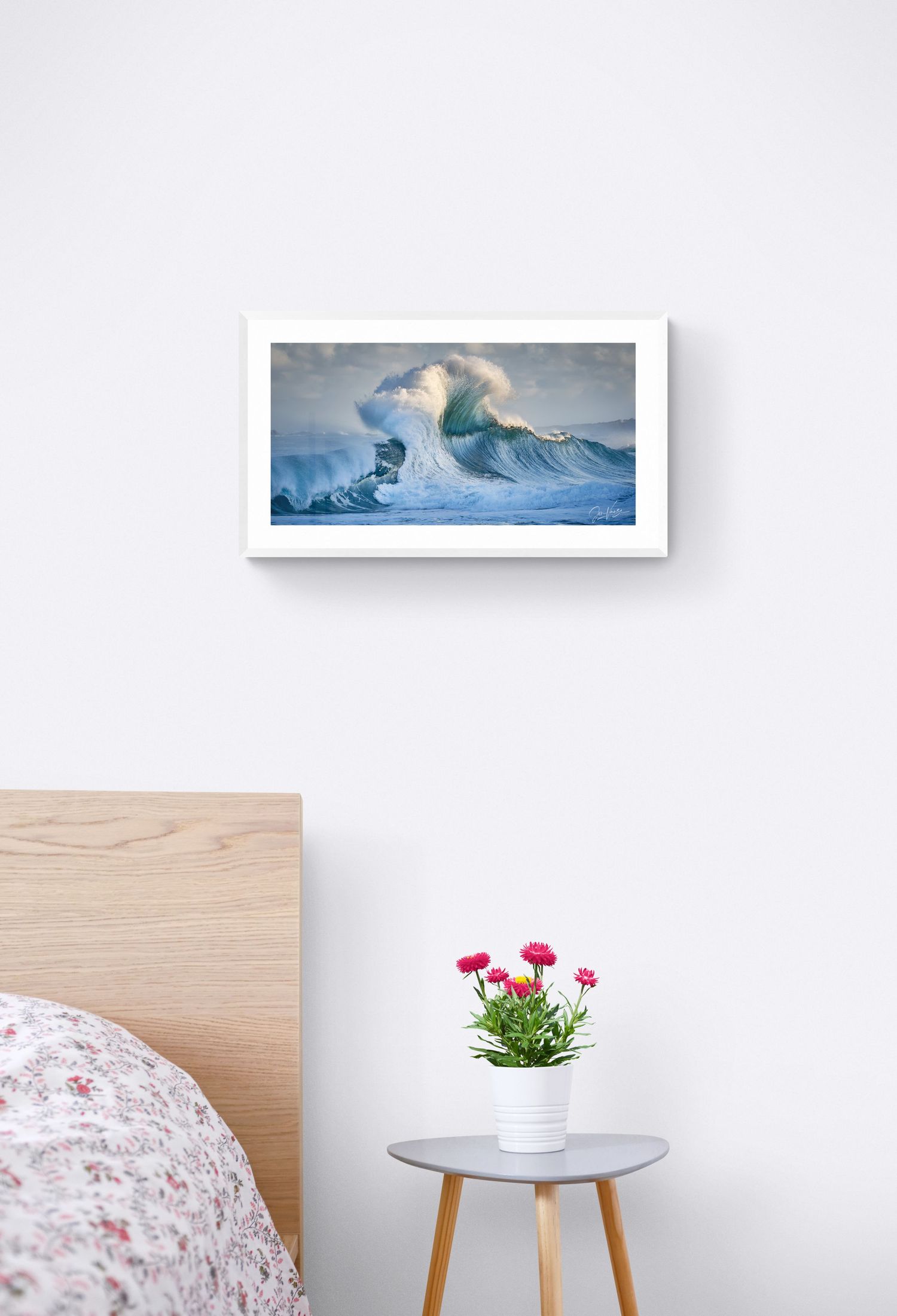 A framed ocean wave painting above a small table with pink flowers in a minimalist bedroom setting.