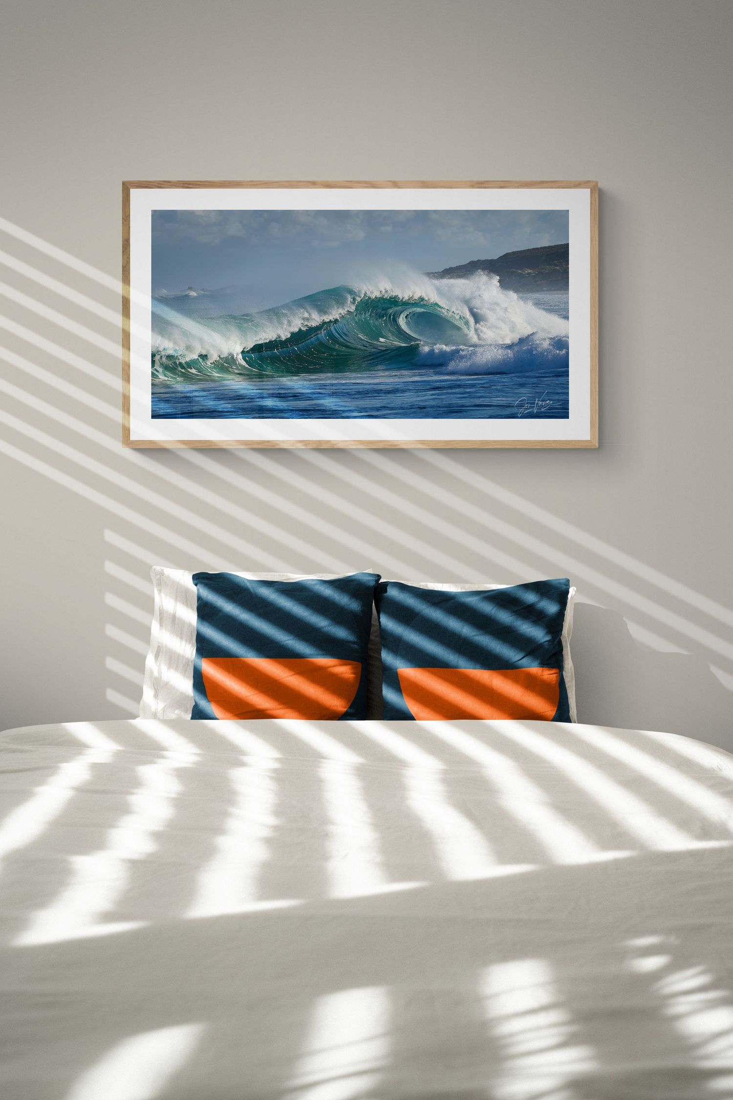 Large wave photograph with blue-green ocean swells mounted above bed with orange and blue striped pillows.