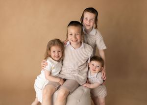 Group portrait of four siblings in matching outfits sitting on light colored chair.