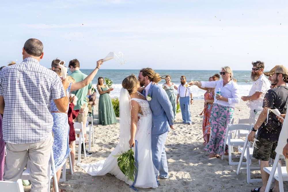 Wedding guests celebrate on beach as couple exchanges vows under bright blue sky.