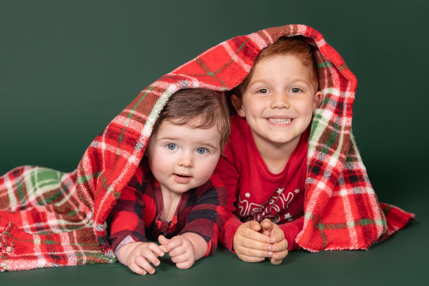 brothers laying on stomach in christmas pajamas with plaid blanket on solid green backdrop in studio clayton new jersey