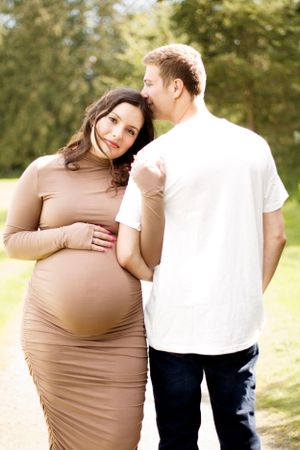 Expecting couple shares an intimate moment outdoors as she wears a fitted tan dress highlighting her pregnancy.