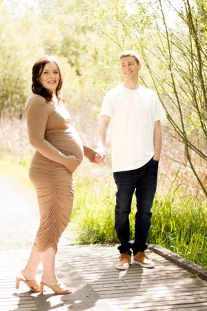 Couple standing on wooden boardwalk during sunny maternity photoshoot in nature.