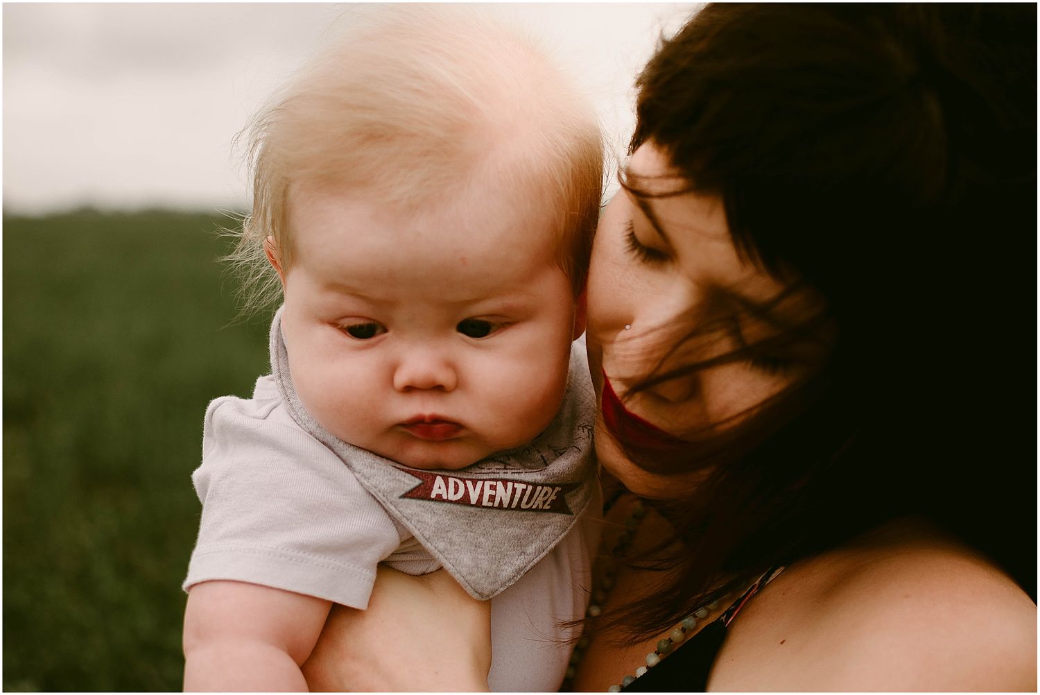 Intimate close-up moment between parent and infant wearing gray shirt in green field.