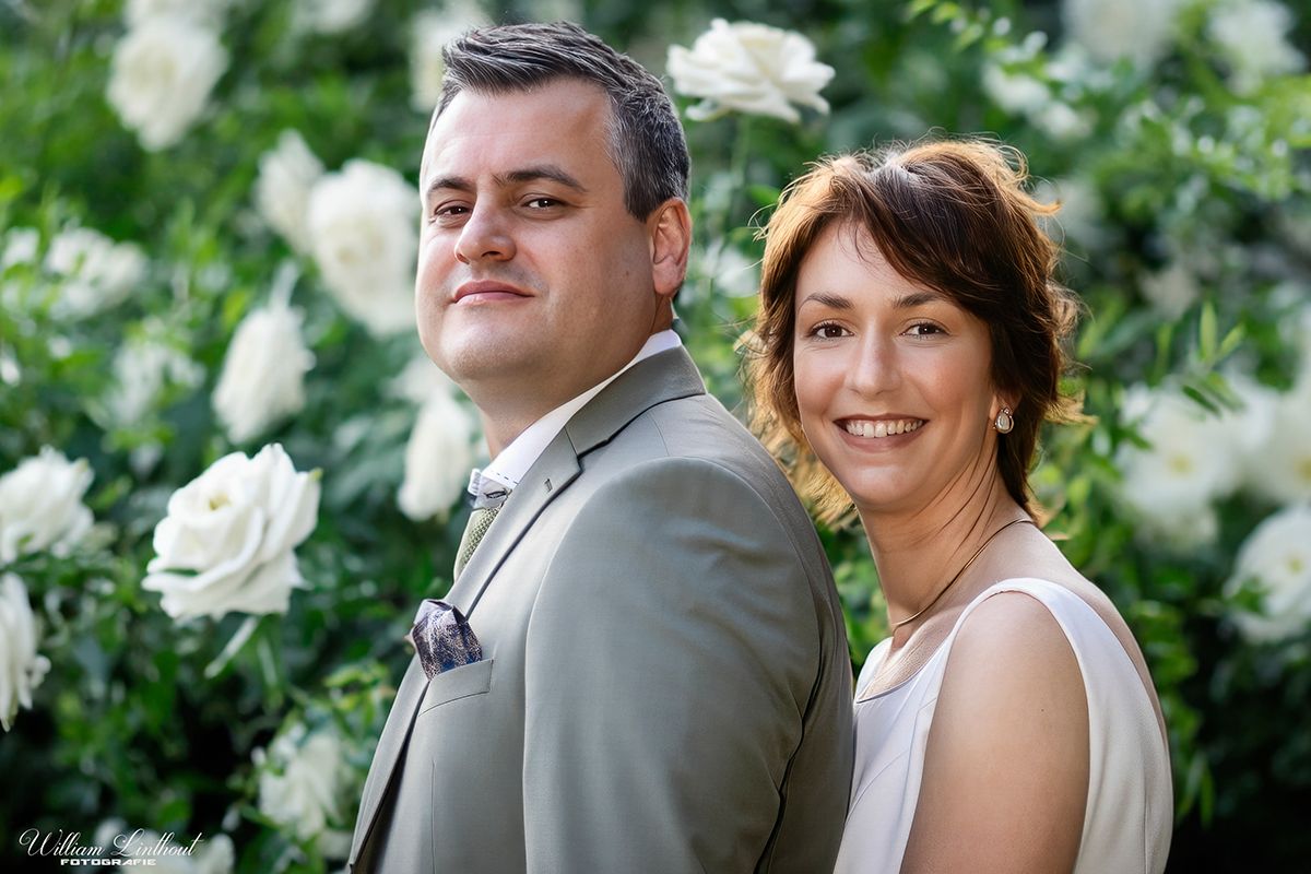 A couple poses together in front of a backdrop of white roses in a garden setting.