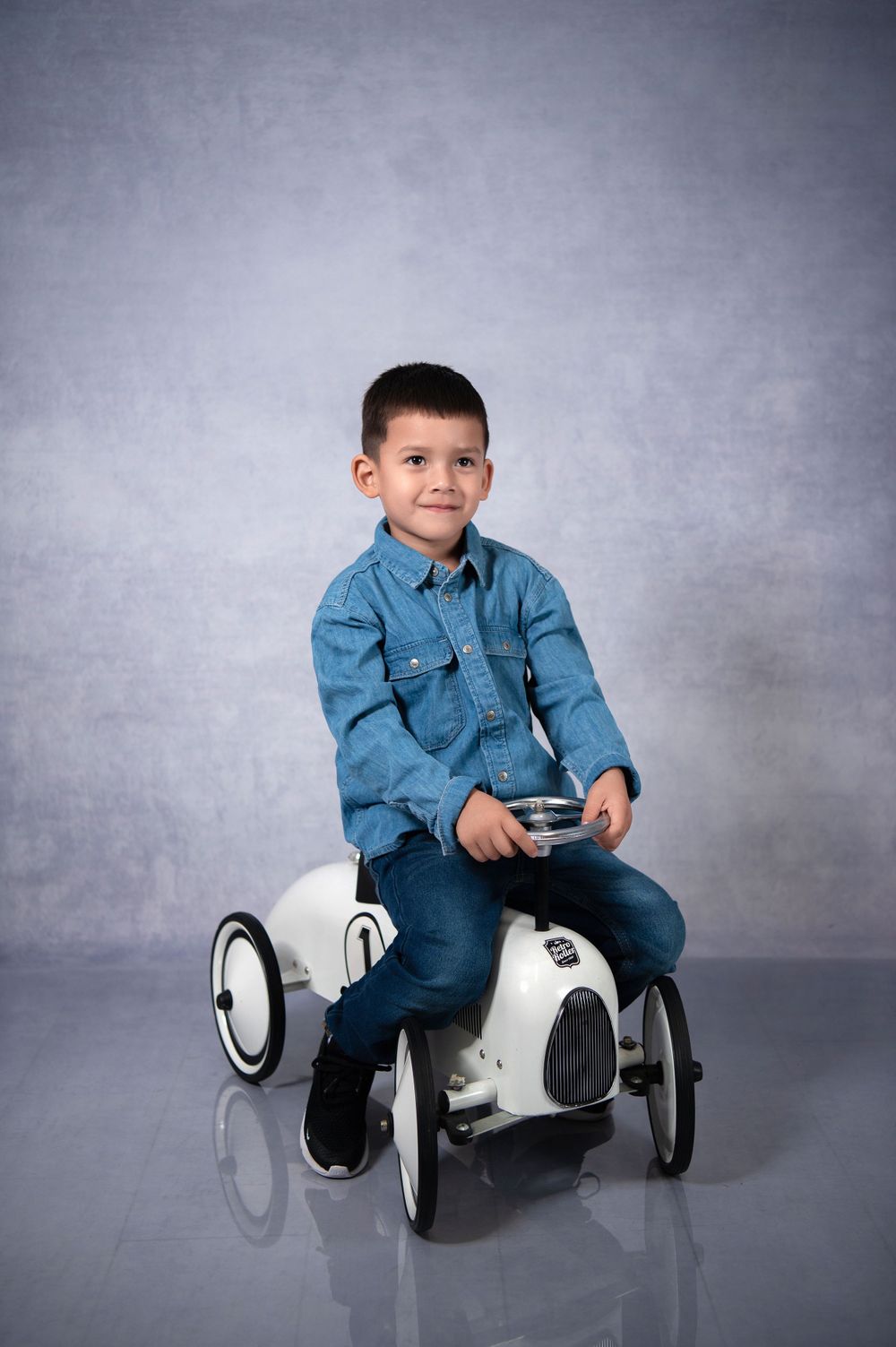 Child riding white vintage-style pedal car in studio setting with grey background.