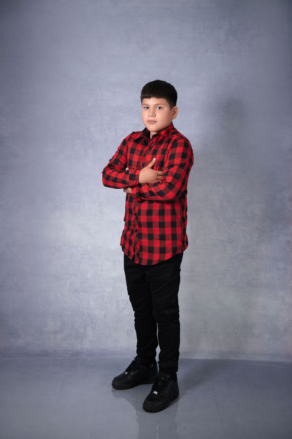 Young child wearing a red and black buffalo plaid shirt and black pants poses against a gray backdrop.