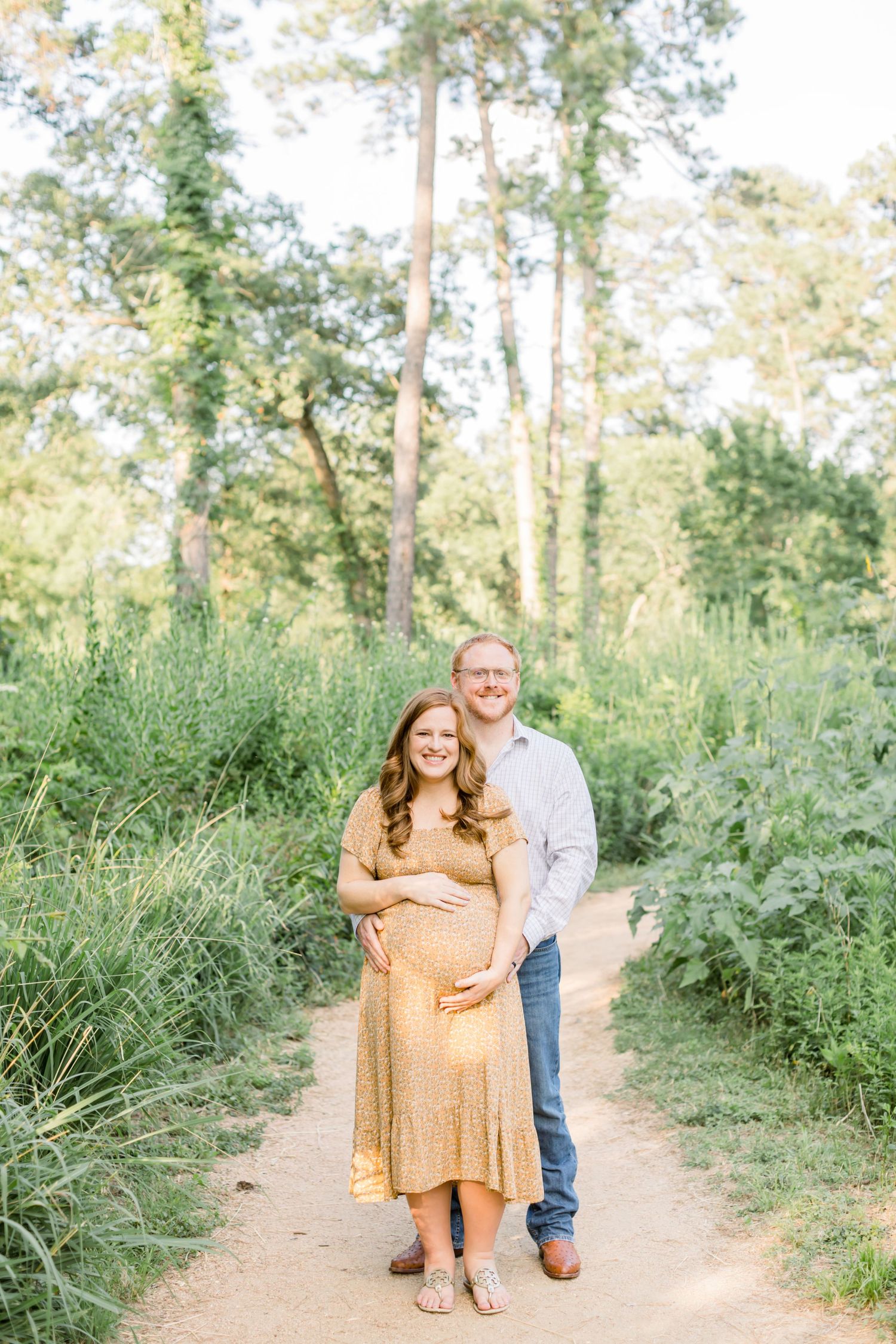 A couple poses together on nature trail surrounded by tall grasses and trees.
