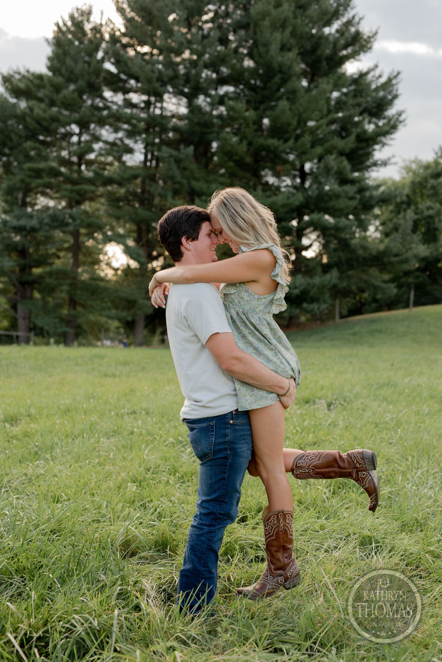 Playful couple embraces in field with pine trees as sun sets.